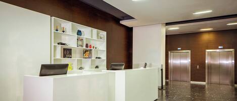 A reception area with a white reception desk, a shelf with various items, and two elevators.