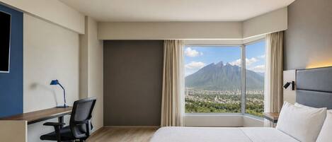 Habitación de hotel con cama, escritorio con silla, lámpara y una ventana grande con vistas a la montaña.