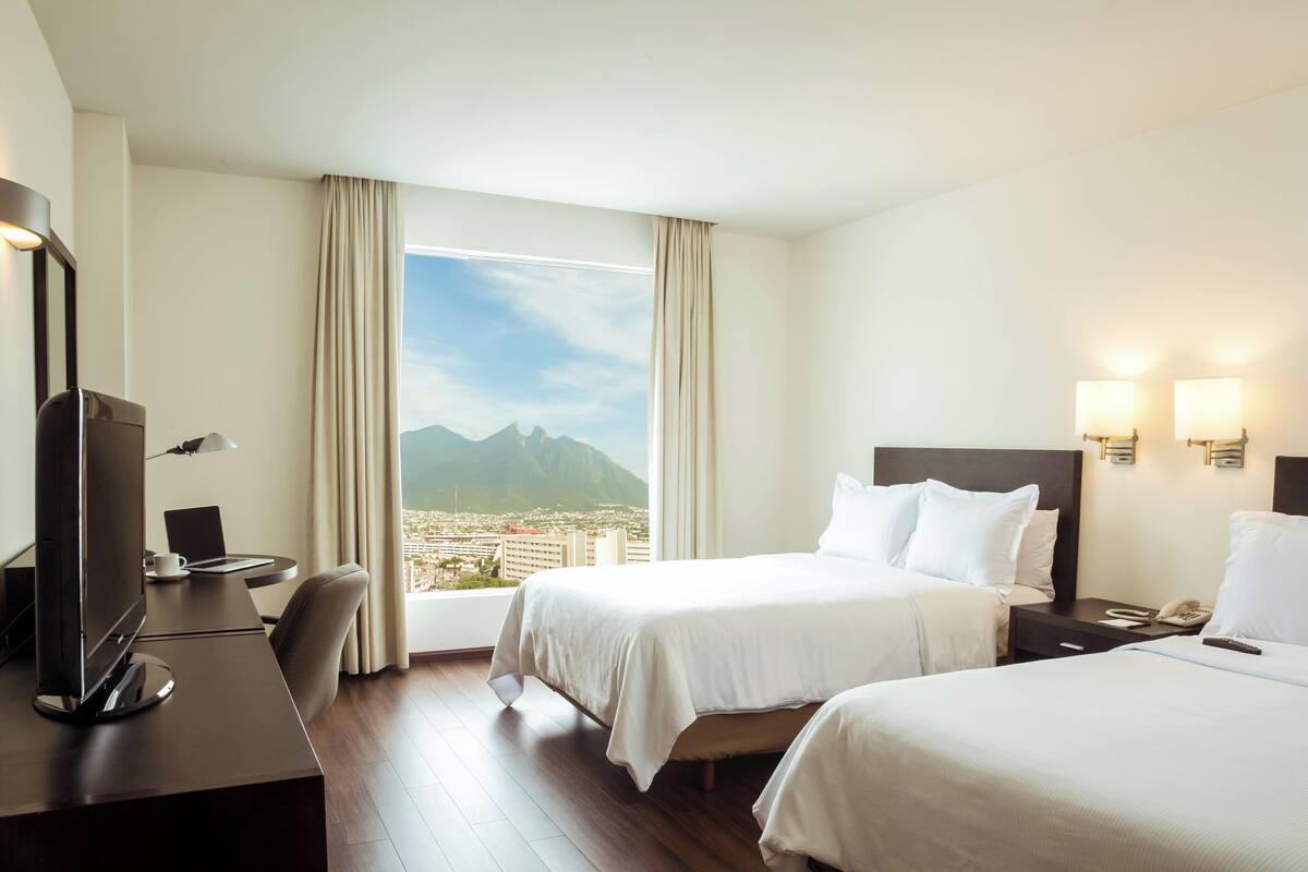 Habitación de hotel con dos camas, un escritorio con televisor, una silla y vistas a la ciudad y a las montañas.