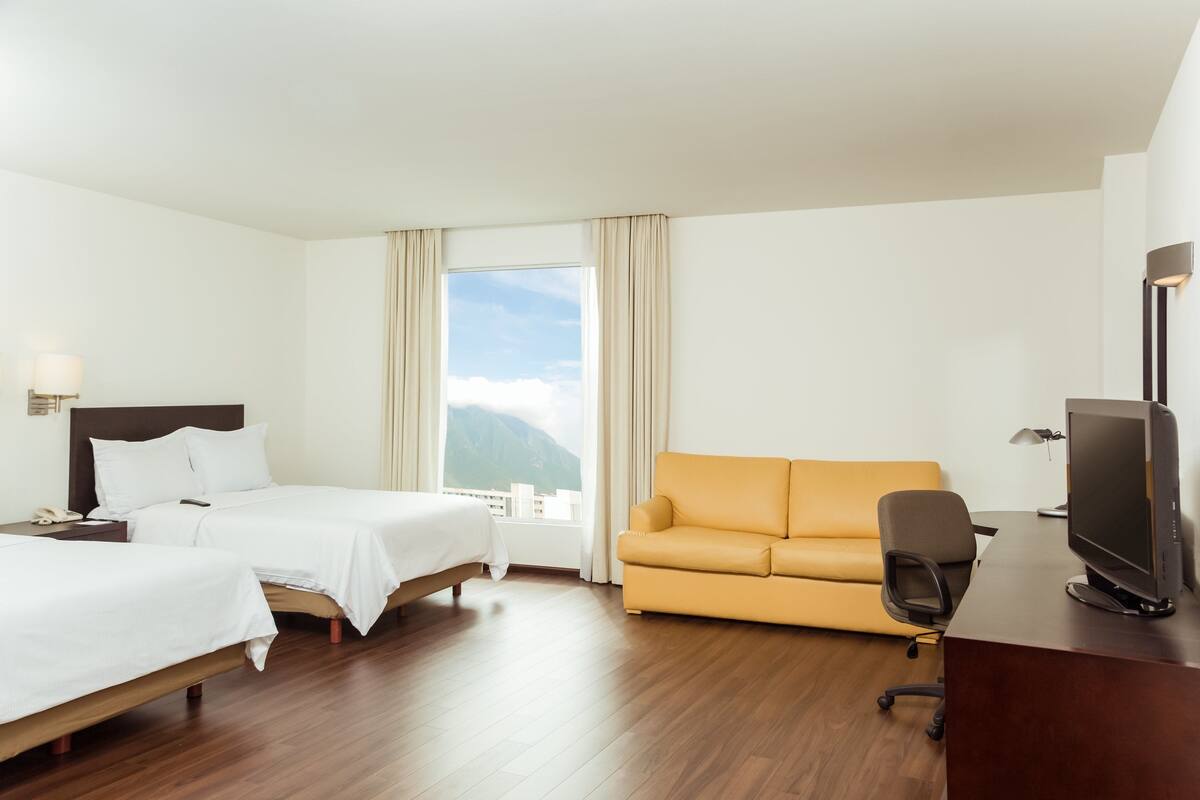 Habitación de hotel con dos camas, un sofá, un escritorio con ordenador y ventana con vistas a la montaña.
