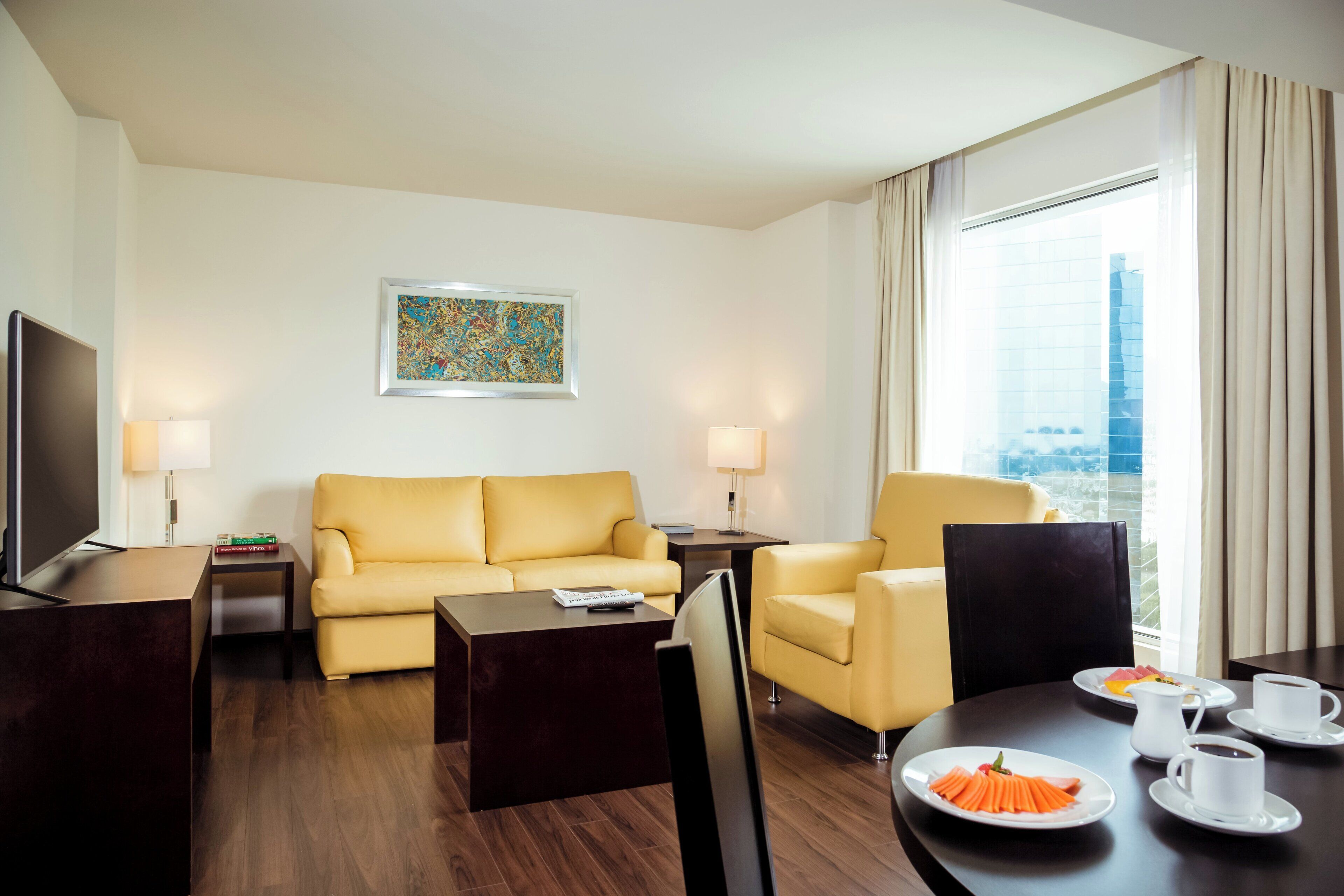 A hotel room with a sofa, armchairs, a dining table with plates of food, a TV, and a window with curtains.