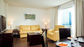 A hotel room with a sofa, armchairs, a dining table with plates of food, a TV, and a window with curtains.