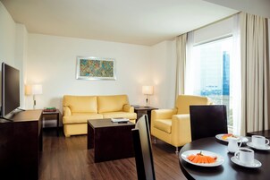 A hotel room with a sofa, armchairs, a dining table with plates of food, a TV, and a window with curtains.