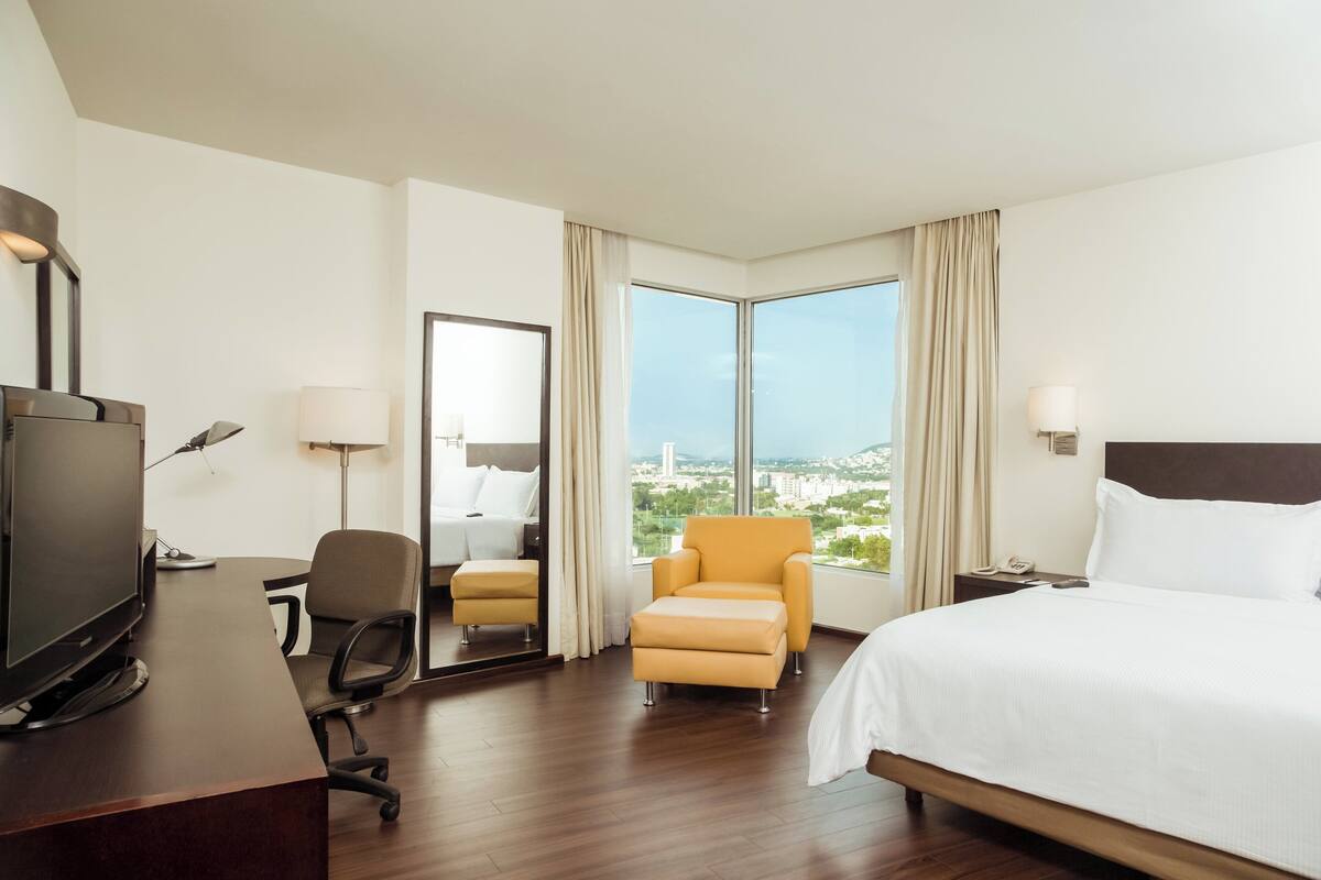 Habitación de hotel con cama, escritorio con televisor, una silla y vista a un paisaje urbano a través de amplias ventanas.