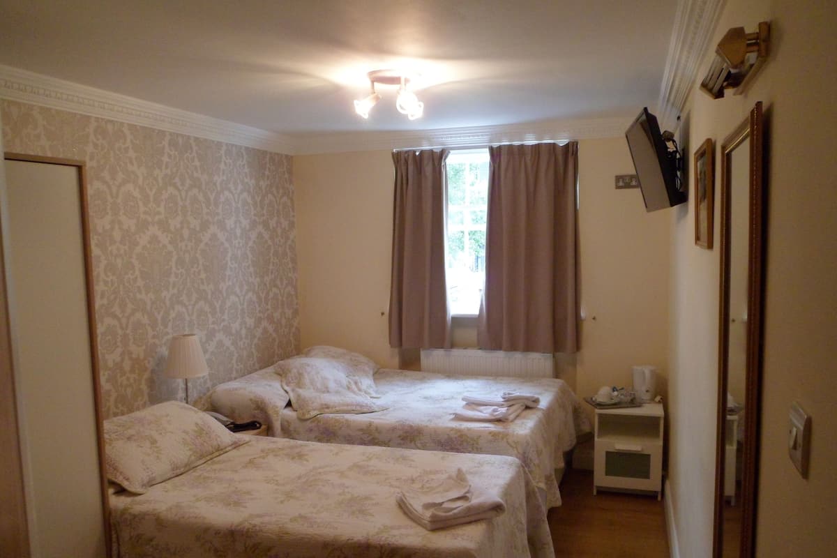 A hotel room with two beds, a nightstand, a lamp, a window with curtains, and a mirror on the wall.