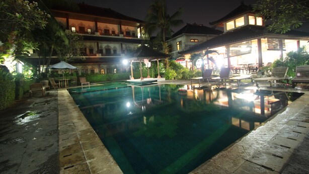 Un área de piscina nocturna con piscina despejada, sillas de descanso y un edificio de varios niveles con terraza.