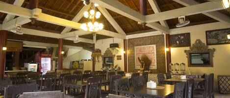 A spacious dining area with high ceilings and wooden beams, featuring multiple tables and chairs, a chandelier, and traditional decor.