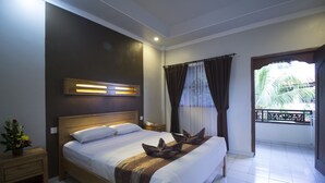 Una cama bien hecha con cabecera de madera, una mesita de noche con lámpara, y vistas a un balcón con una palmera.