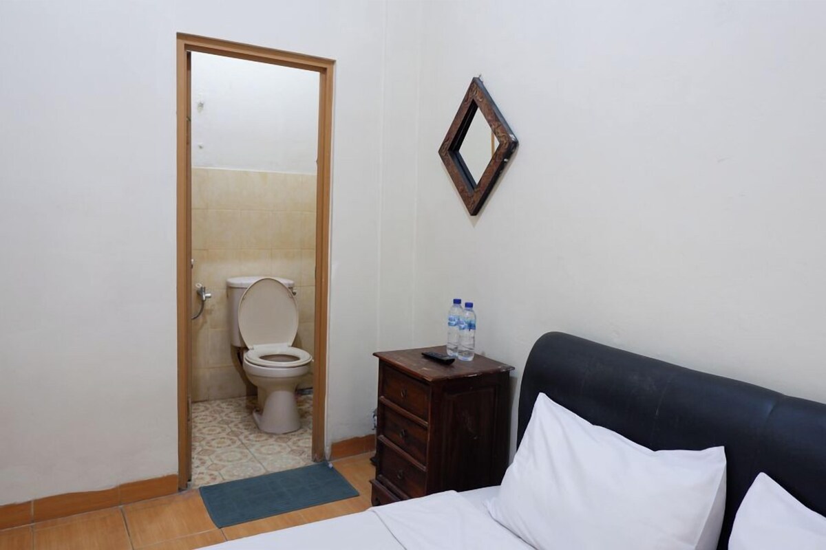 Una habitación pequeña y limpia con una cama, un aparador de madera, un espejo y un inodoro visible a través de una puerta abierta.