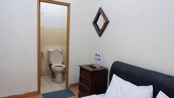 Une petite pièce propre, avec un lit, une commode en bois, un miroir et des toilettes visibles à travers une porte ouverte.