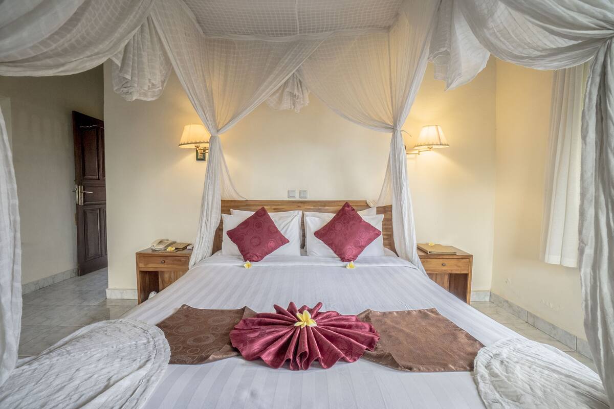 Un lit à baldaquin avec du linge de lit blanc, deux oreillers rouges et un élément décoratif central aux couleurs rouge et marron.