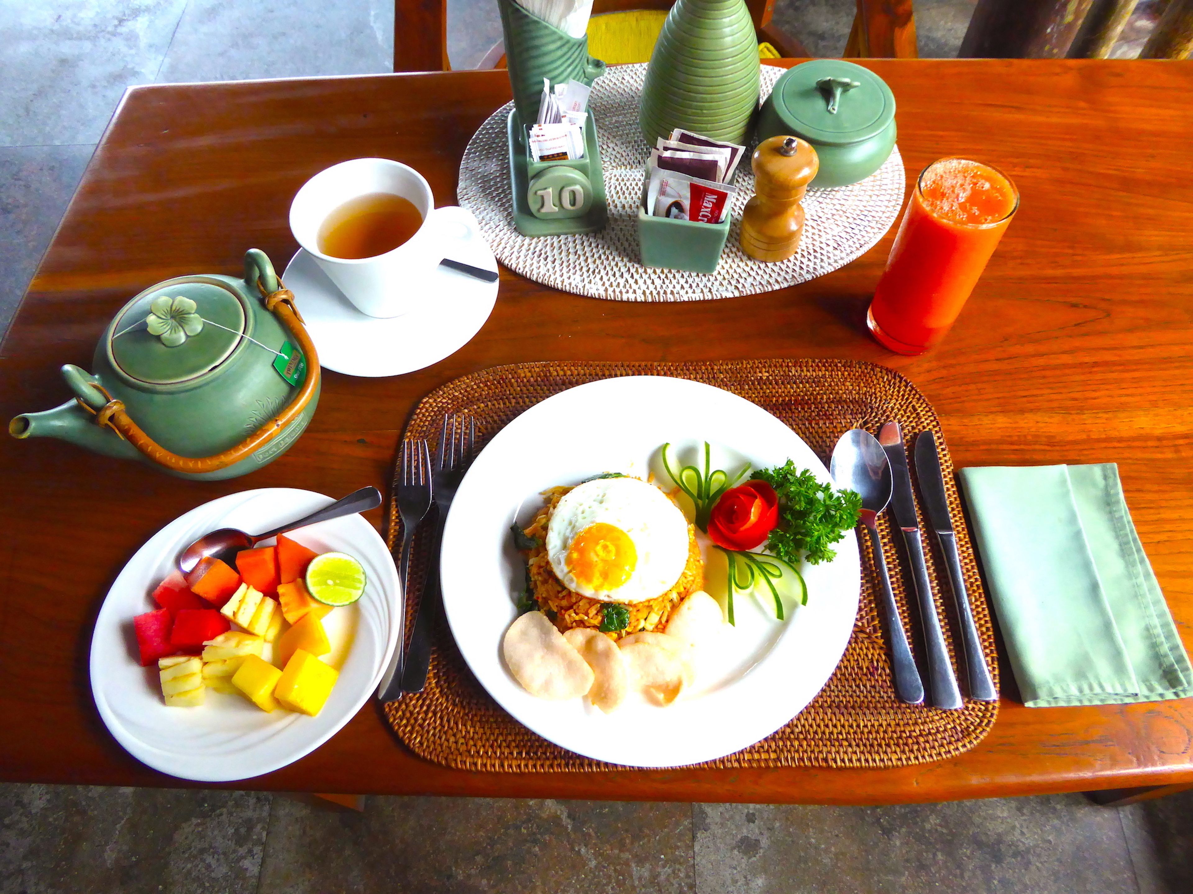 A table set with a teapot, cups, a fruit bowl, a plate with eggs, vegetables, and a glass of juice.