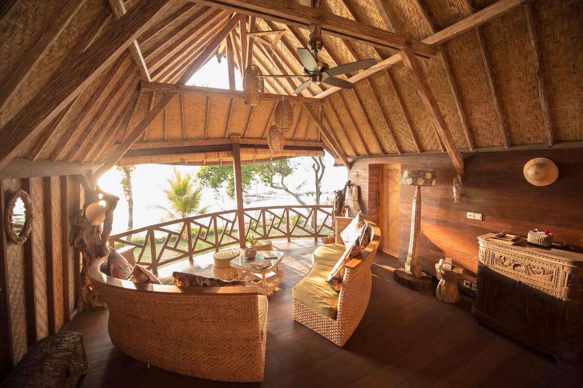 Un intérieur caractérisé par un plafond haut, des poutres en bois, des meubles en osier et une vue accessible depuis un balcon.