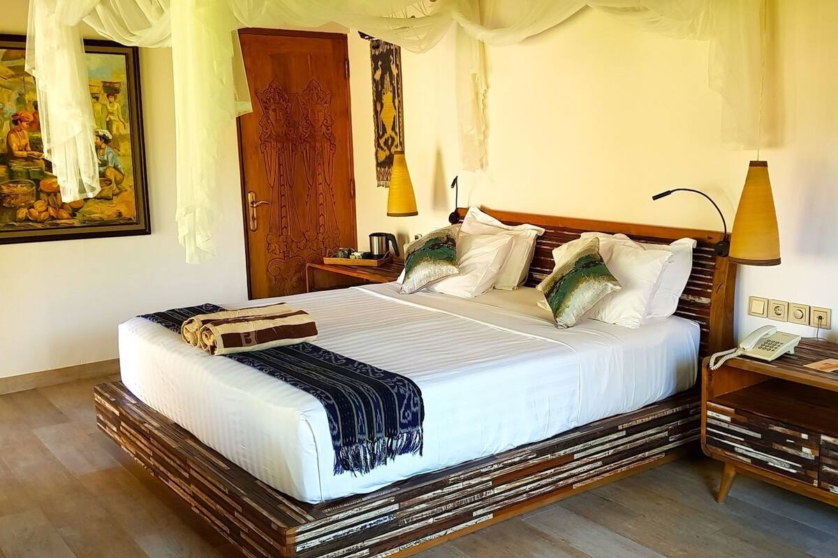 Un lit avec un cadre en bois, une literie blanche et des coussins décoratifs. Il y a une table de chevet avec une lampe, un téléphone et des bocaux décoratifs. Un tableau est accroché au mur au-dessus du lit.