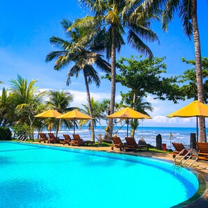 Une piscine entourée de chaises longues et de parasols, bordée de palmiers et offrant une vue sur l’océan.