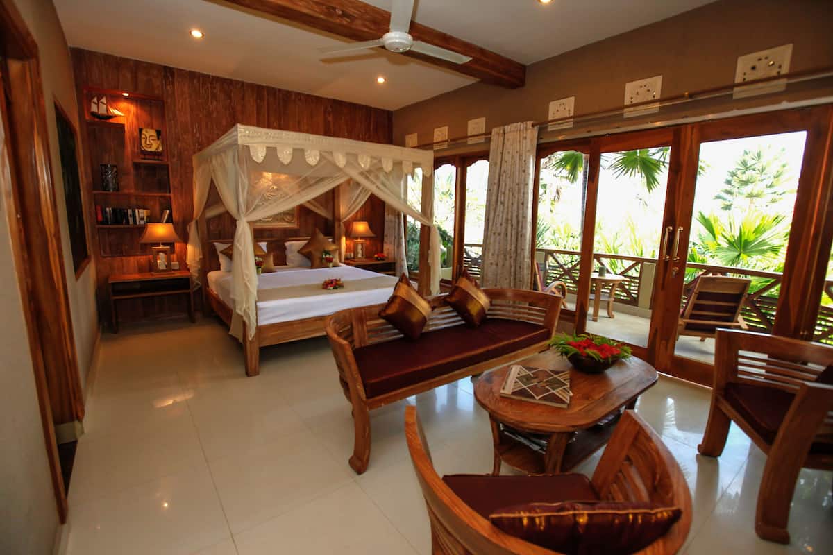Une chambre avec un lit à baldaquin, un coin salon avec une table et des chaises, et un balcon aménagé avec du mobilier d’extérieur.