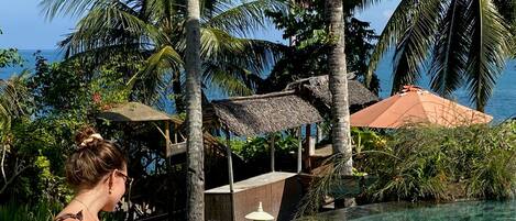 A couple relaxing in a clear pool with a wicker tray of drinks and snacks, surrounded by palm trees and a thatched-roof structure.