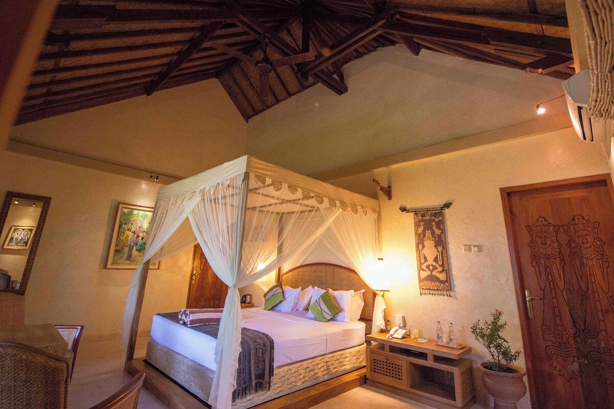 A room with a four-poster bed, a nightstand with a lamp, a wooden door with carvings, and a wall-mounted tapestry.