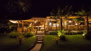 A well-lit outdoor area with a wooden structure, palm trees, and a pathway leading to a building. - Puri Dajuma Beach Eco-Resort & Spa (Pekutatan)