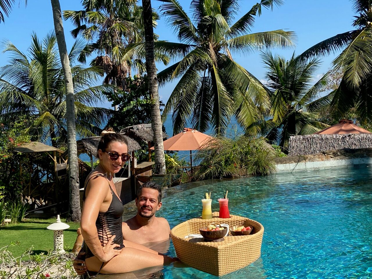 Una pareja relajándose junto a una piscina con una mesa de mimbre que sostiene bebidas y aperitivos, rodeada de palmeras y un cielo azul despejado.