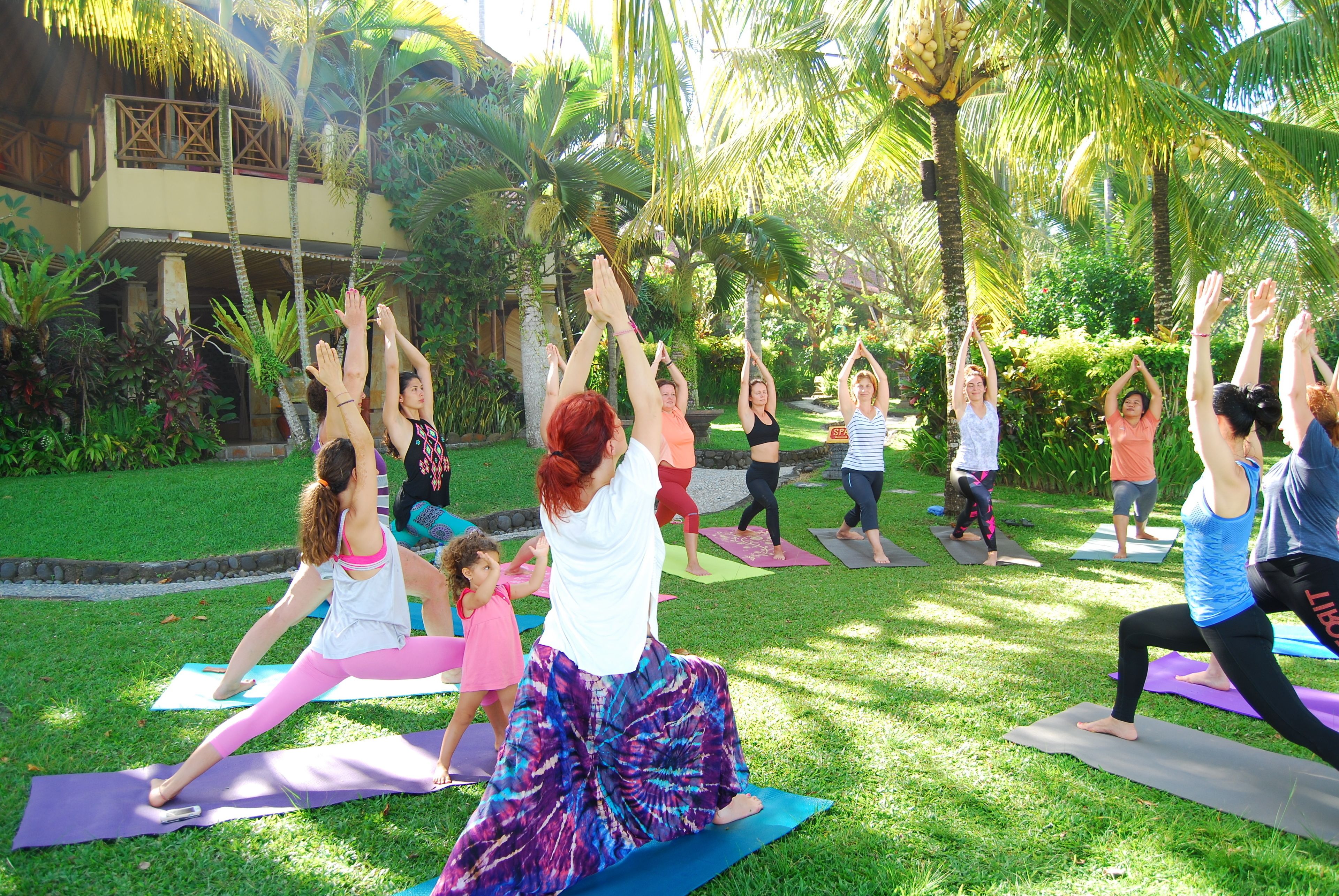 Un grupo de personas practicando yoga sobre esterillas en un entorno al aire libre con palmeras y un edificio de fondo.