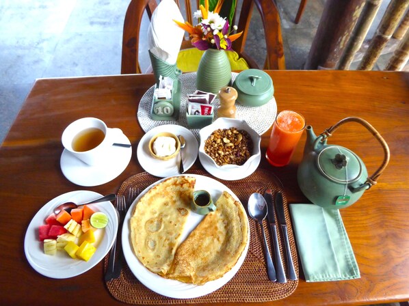 Una mesa de desayuno con una fuente de fruta, un tazón de granola, una taza de café, una tetera y un vaso de jugo.