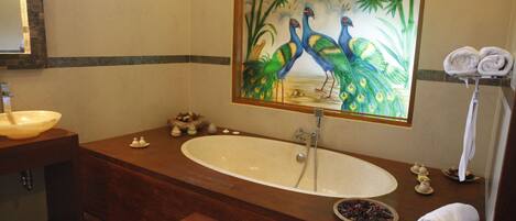 A bathroom with a large oval sink, a wooden vanity, a peacock painting above the mirror, and a towel rack with folded towels.