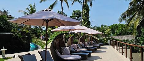 A row of sun loungers with umbrellas, a pool, and palm trees in the background.