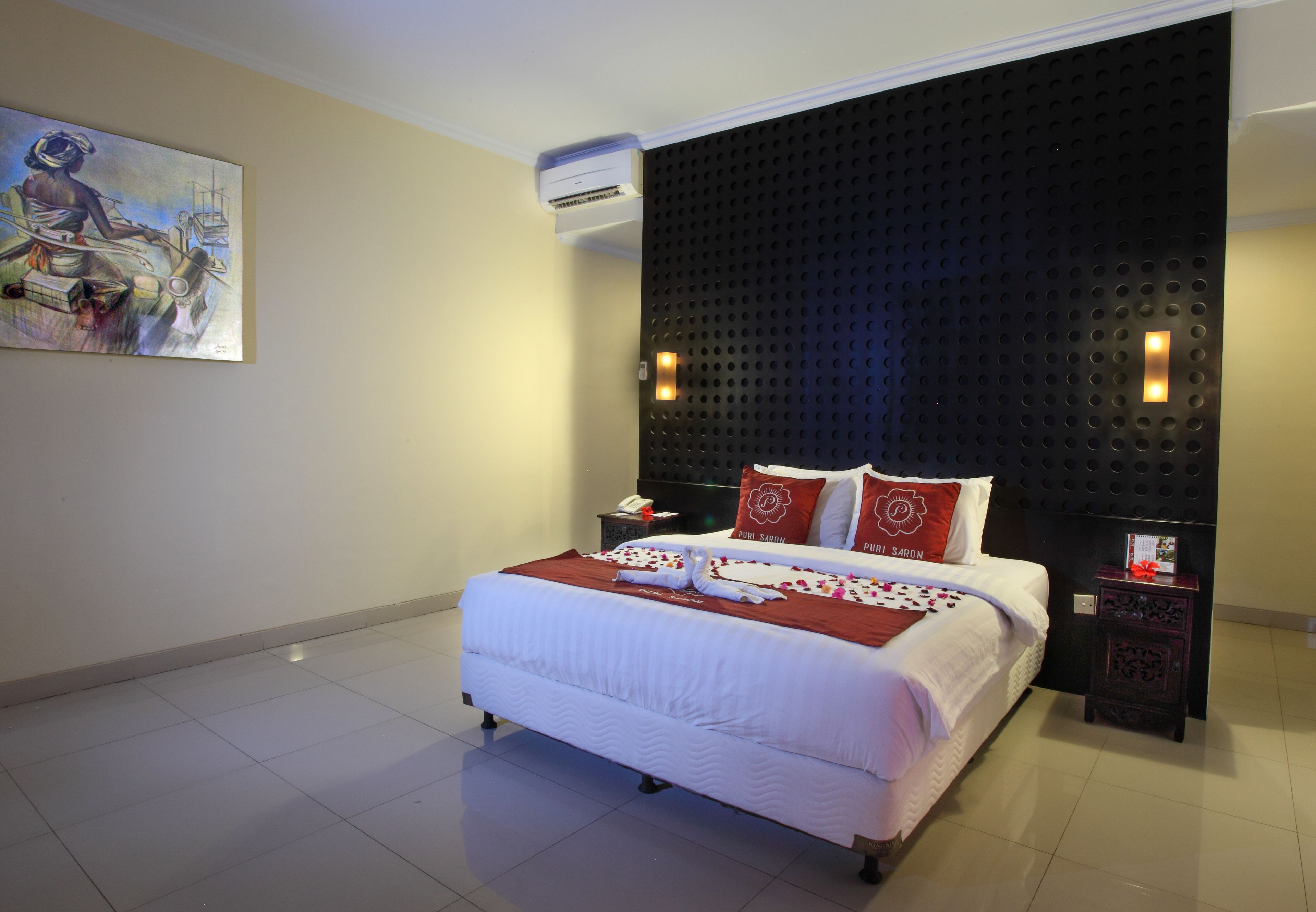 Una cama bien tendida con sábanas blancas y almohadas rojas, un cuadro en la pared y un cabecero negro con textura.