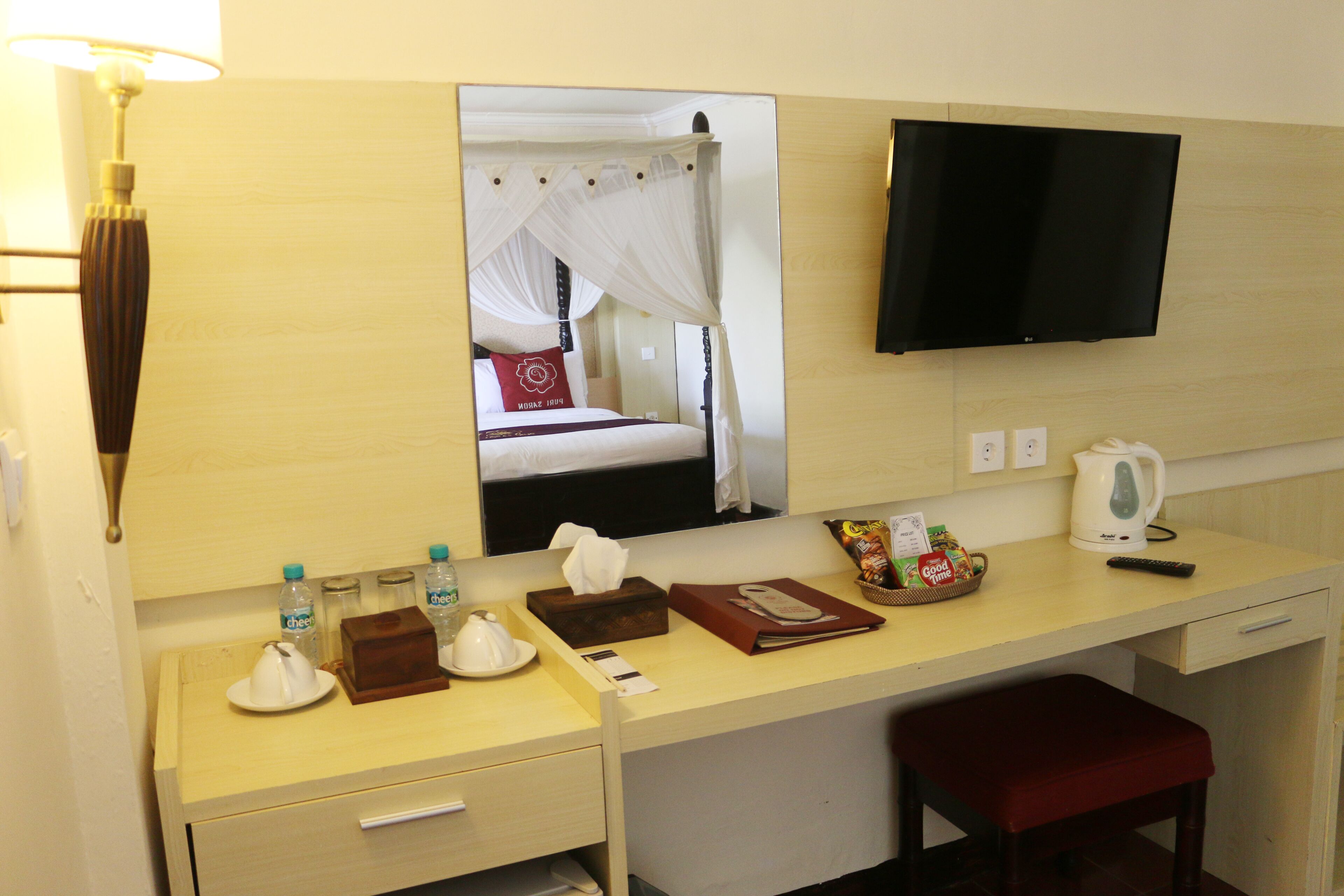 Habitación de hotel con cama, escritorio, televisor, espejo, mesita de noche, una silla roja y ventana con cortinas.