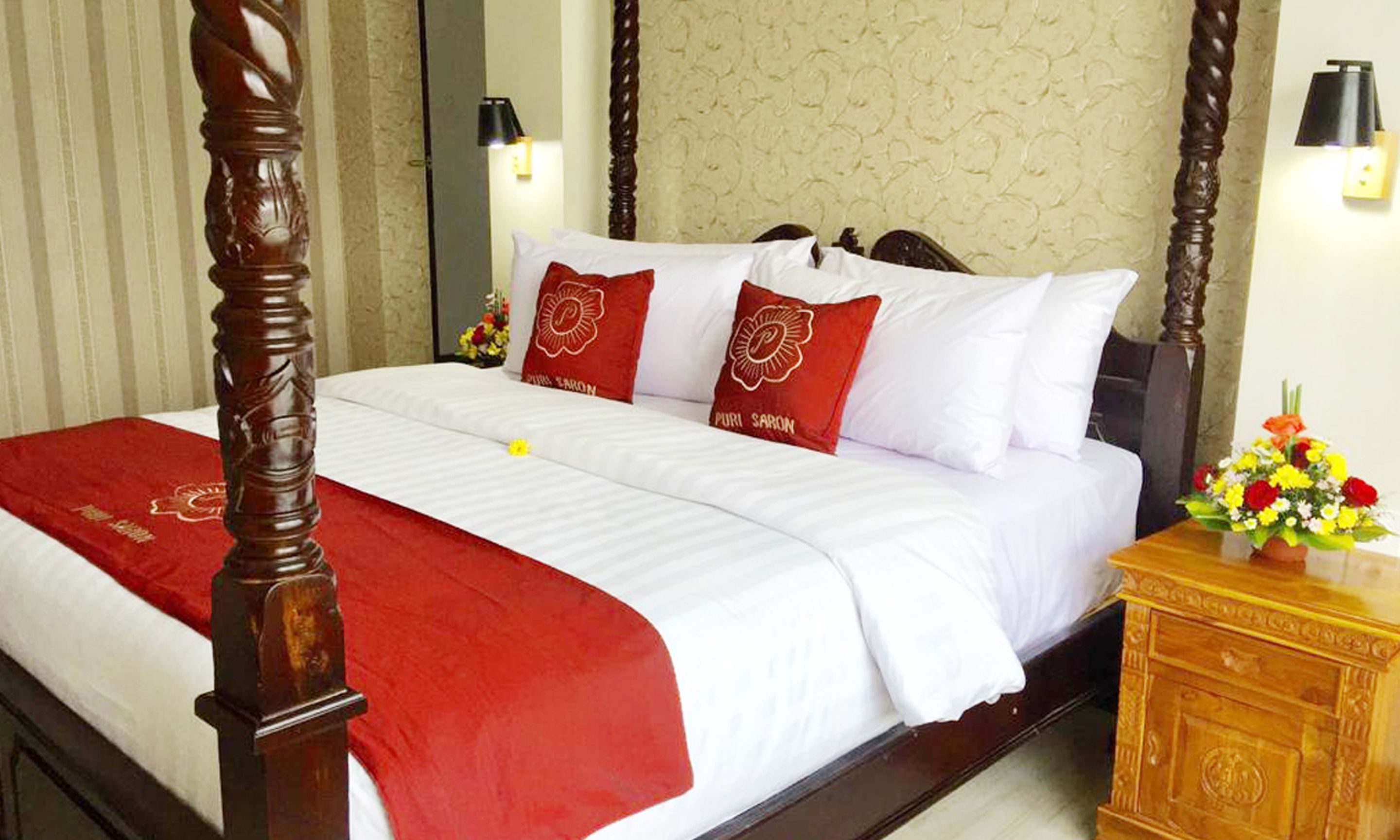 Una habitación de hotel con cama con dosel, ropa de cama blanca, almohadas decorativas rojas y una mesita de noche de madera con un jarrón de flores.