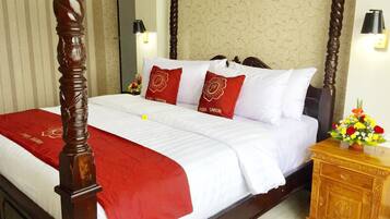Una habitación de hotel con cama con dosel, ropa de cama blanca, almohadas decorativas rojas y una mesita de noche de madera con un jarrón de flores.