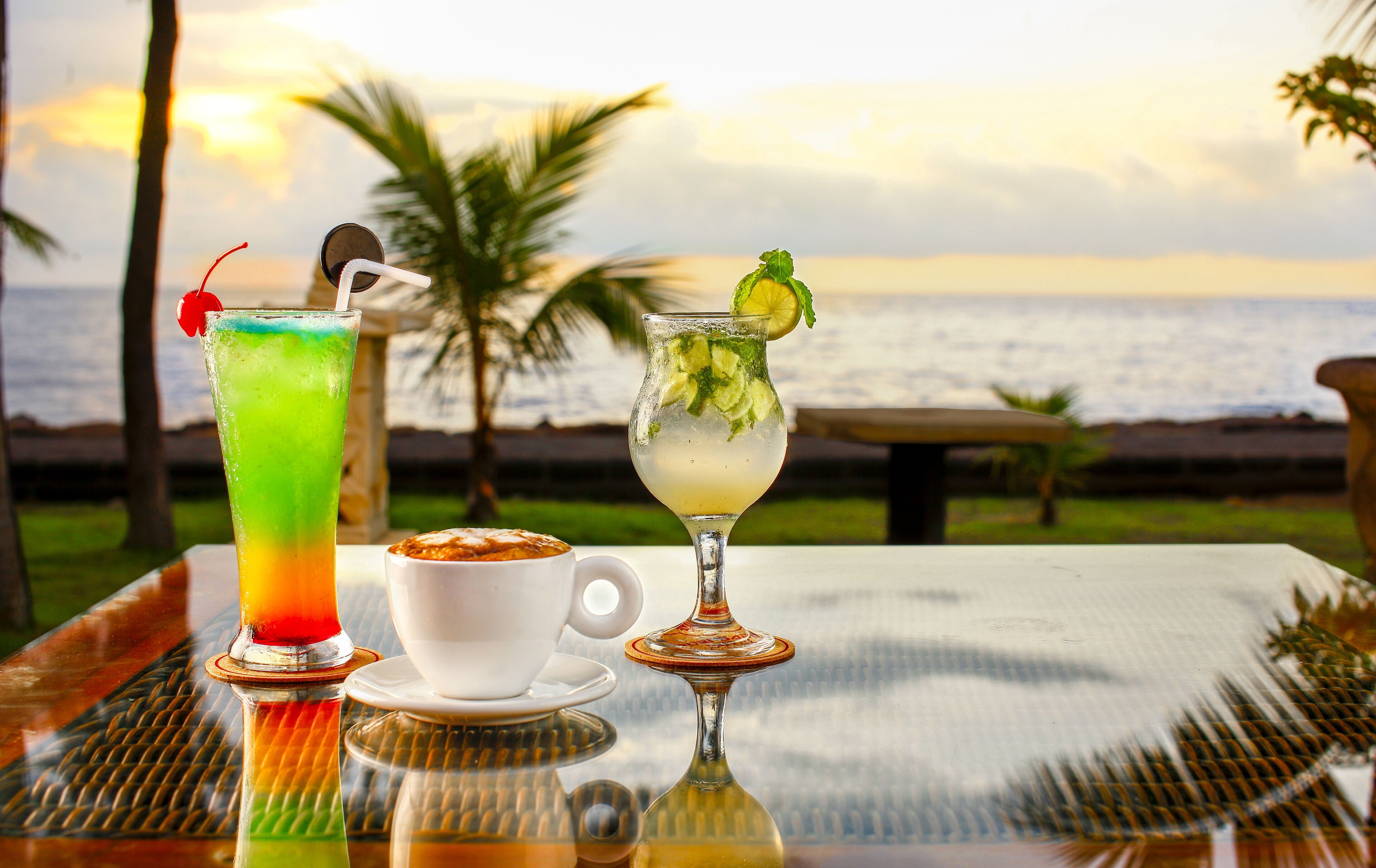 Two cocktails and a cup of coffee on a table with a view of the ocean and palm trees.