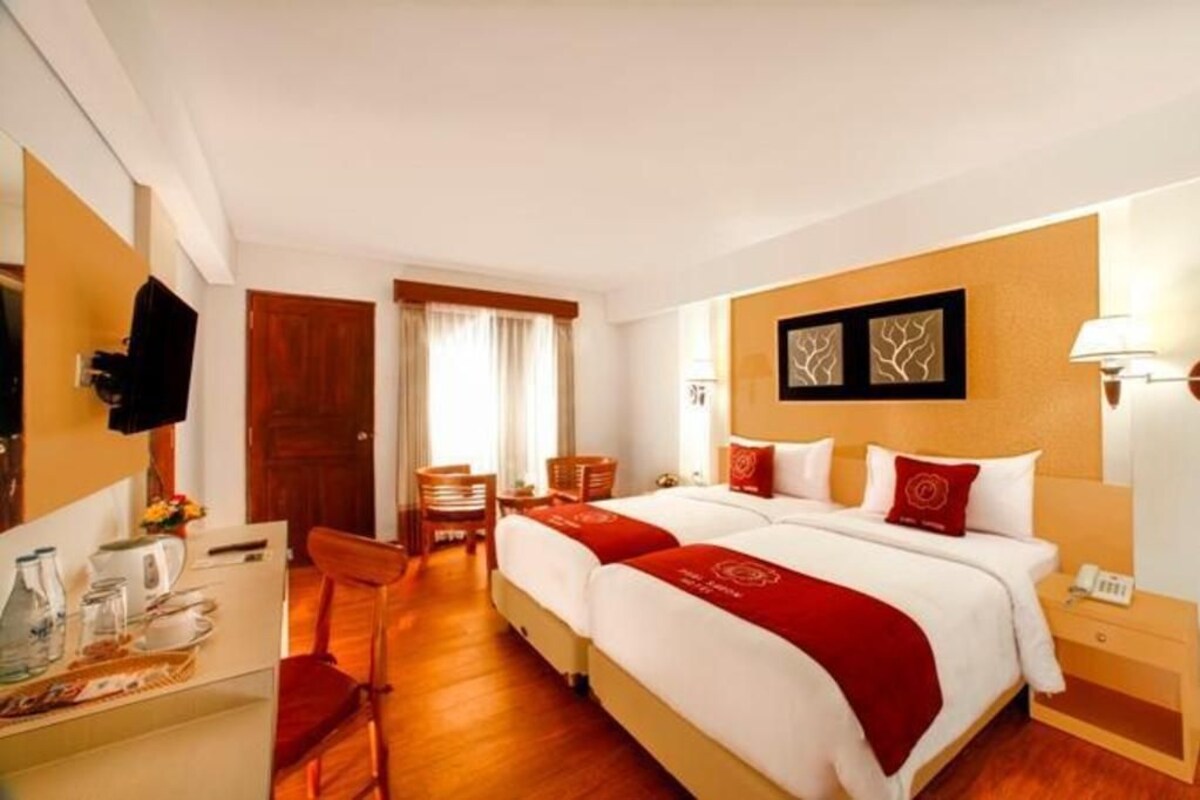 A hotel room with two beds, a desk with a chair, a small table with a lamp, and a door leading to another room.