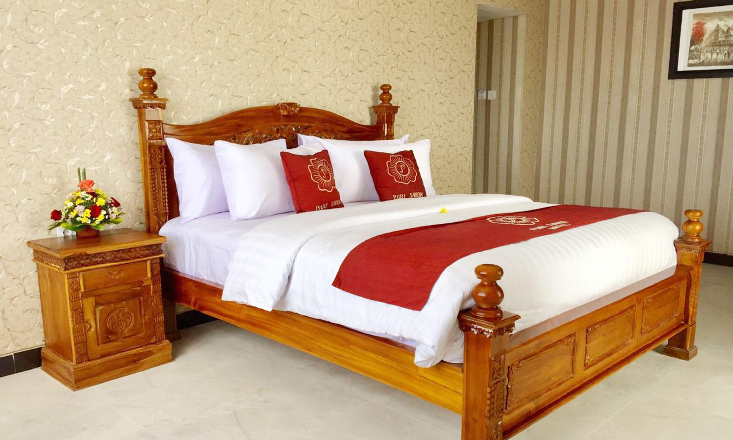 A wooden four-poster bed with white and red bedding, a nightstand with a vase of flowers, and a striped wallpapered wall.