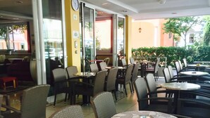 An outdoor cafe with wicker chairs and round tables, featuring a glass door entrance and a clock on the wall. - Monica (Rimini)