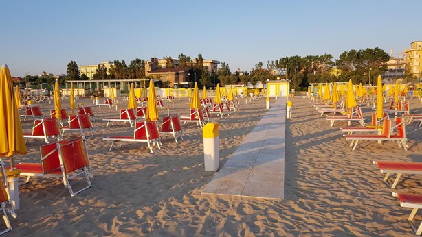 A beach with rows of red and yellow sun loungers and umbrellas, a clear sky, and buildings in the background. - Euromar (Rimini)