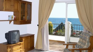 Ein Wohnzimmer mit Holzschrank, Fernseher und einem Sessel. Ein Balkon bietet Blick auf das Meer.