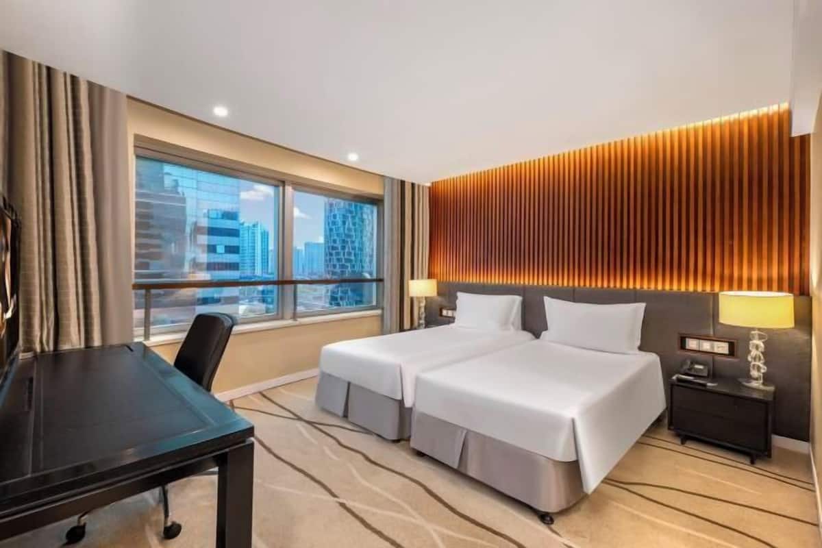 Ein Hotelzimmer mit zwei Betten, einem Schreibtisch, einem Fenster mit Blick auf die Stadt und einer Wand mit vertikalen Holzlatten.