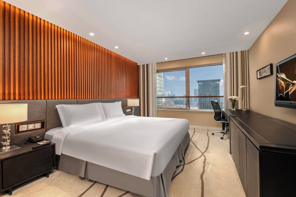 Ein Hotelzimmer mit Bett, Nachttisch, Schreibtisch, Stuhl und einem großen Fenster mit Blick auf die Stadt.