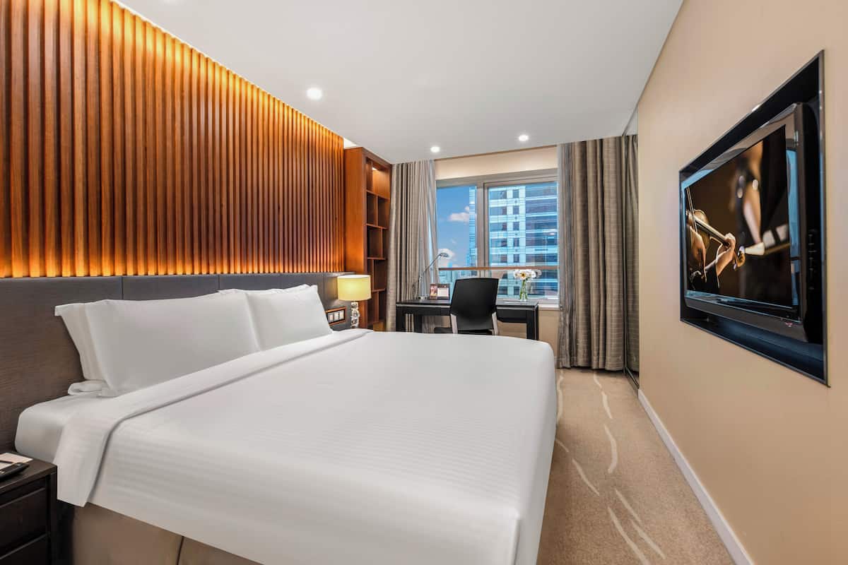 Ein Hotelzimmer mit einem großen Bett, einem Schreibtisch mit Stuhl, einem an der Wand montierten Fernseher und einem Fenster mit Vorhängen.