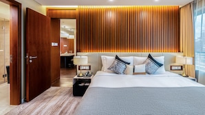 Una camera d'albergo moderna con un letto grande, una testiera in legno, comodini con lampade e vista sulla città attraverso ampie finestre.