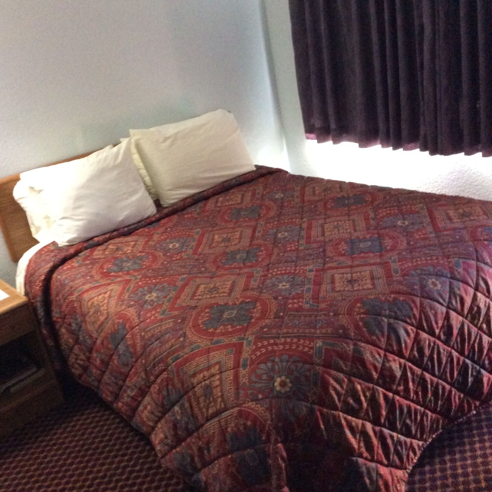 A bed with a patterned quilt, a white pillow, and a dark curtain.