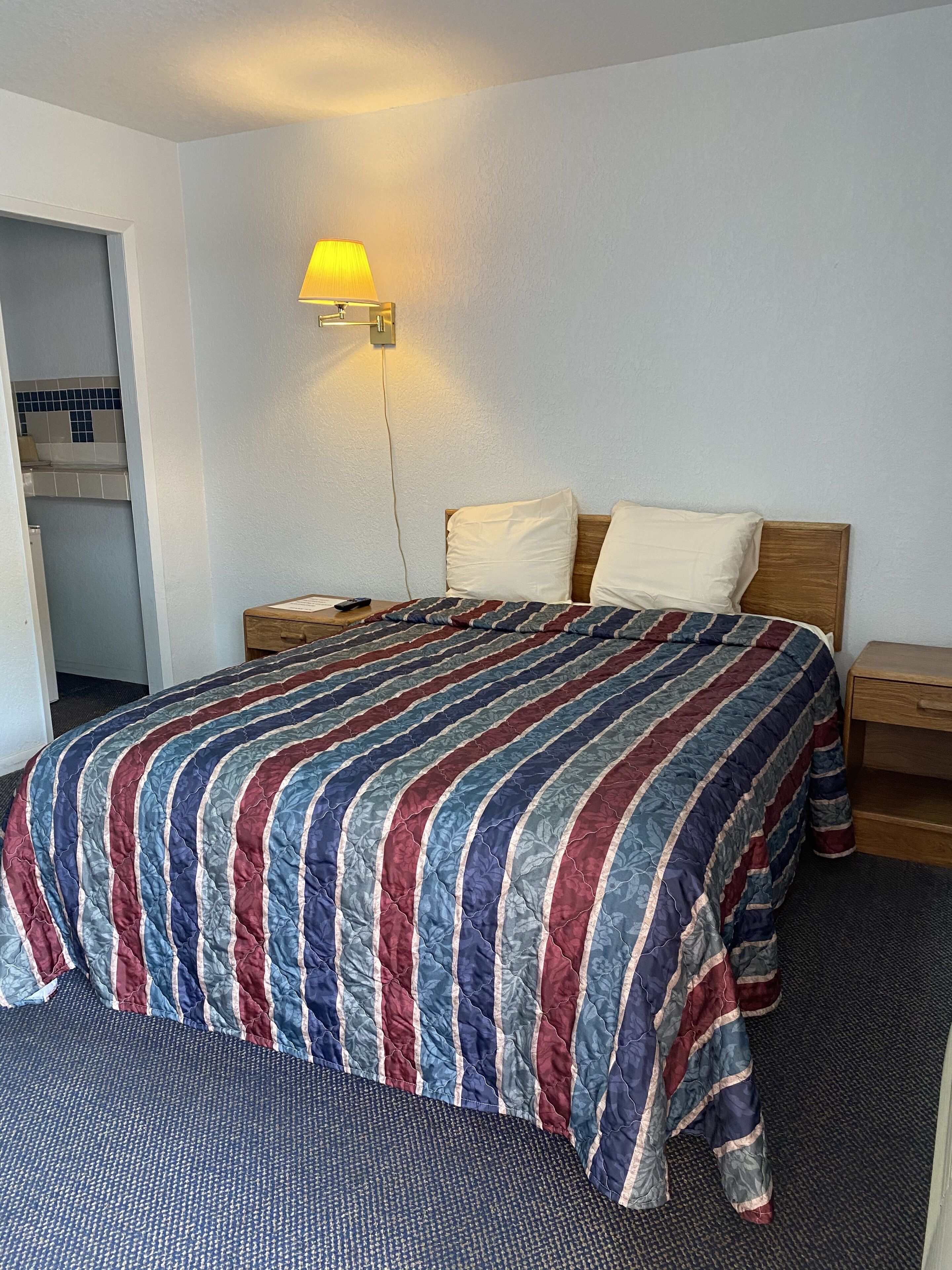 A hotel room with a bed, a nightstand, a lamp, and a view of a kitchen area.