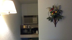 Habitación de hotel compacta con kitchenette, un pequeño refrigerador, microondas, una silla, una mesa con mantel individual y un arreglo floral de pared.