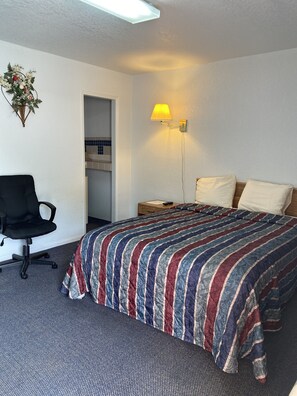Quarto de hotel com cama, escrivaninha, cadeira, mesa de cabeceira e luminária na parede.