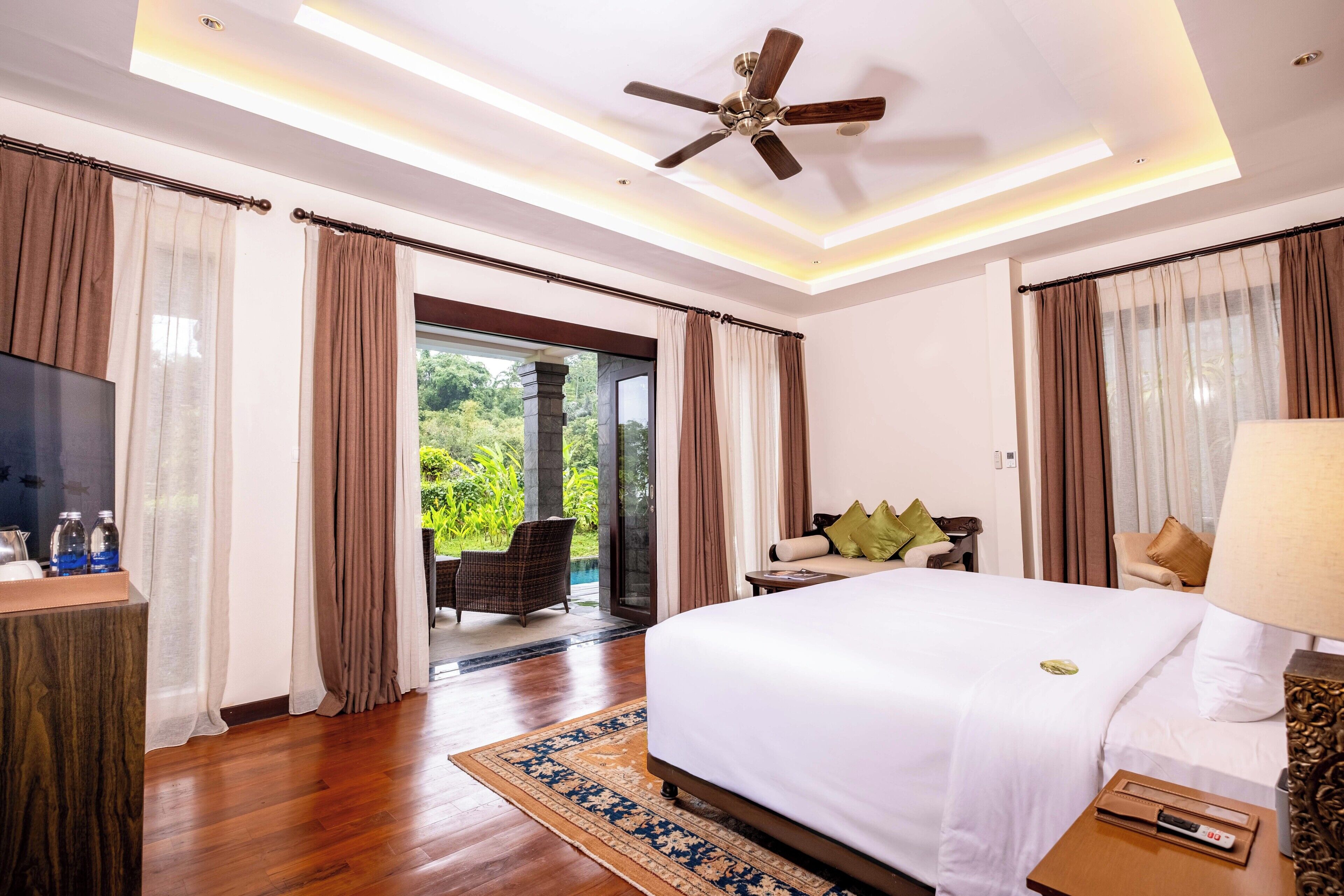 A hotel room with a large bed, a ceiling fan, a TV, and a view of a garden through the sliding glass door.