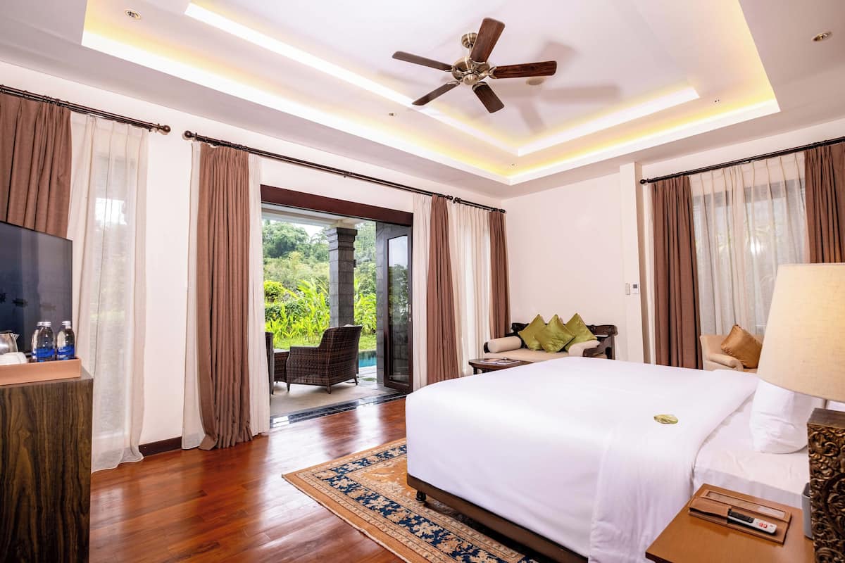 A hotel room with a large bed, a ceiling fan, a TV, and a view of a garden through the sliding glass door.