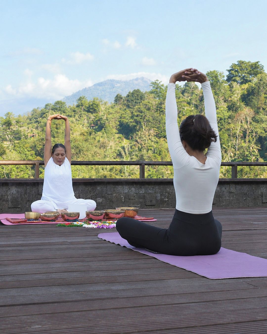 Duas pessoas praticando yoga em um deck de madeira, com montanhas ao fundo.