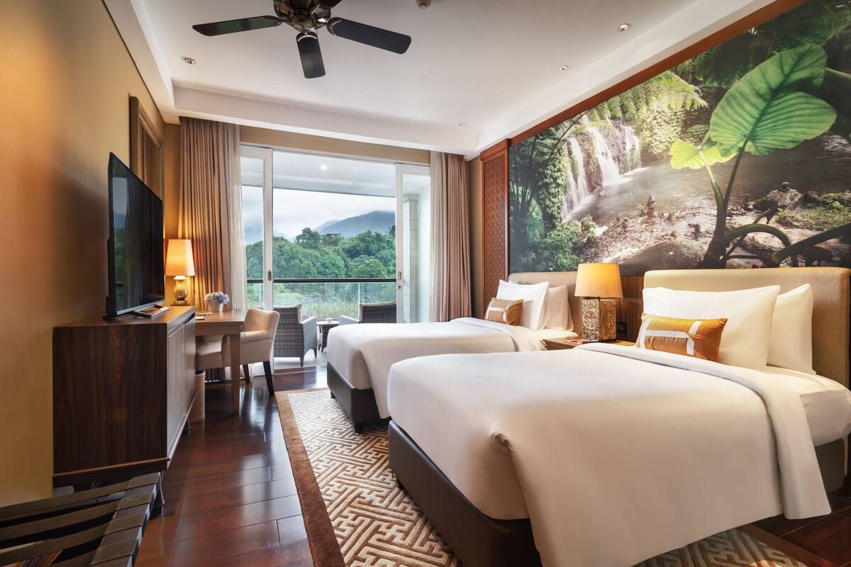 A hotel room with two beds, a desk, a TV, and a large window with a view of greenery.